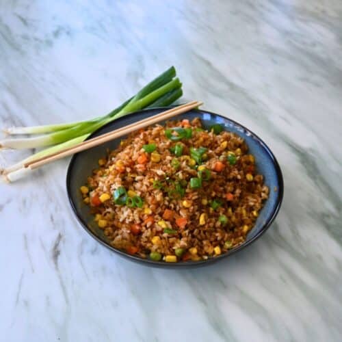 A bowl of air fryer fried rice with mixed vegetables and green onion garnish sits on marble, next to chopsticks and fresh green onions.