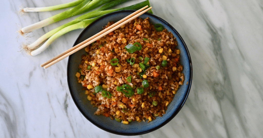 A bowl of air fryer fried rice with colorful vegetables and chopped green onions, with chopsticks and fresh green onions on marble.