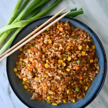 A bowl of air fryer fried rice with corn, peas, and carrots sits on a marble counter, garnished with chopsticks and green onions.