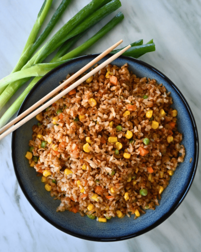 A bowl of air fryer fried rice with corn, peas, and carrots sits on a marble counter, garnished with chopsticks and green onions.