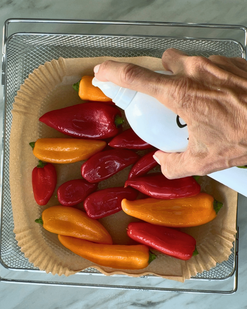 A hand sprays oil onto red and orange mini bell peppers and baby potatoes arranged on parchment in a metal tray for air frying.