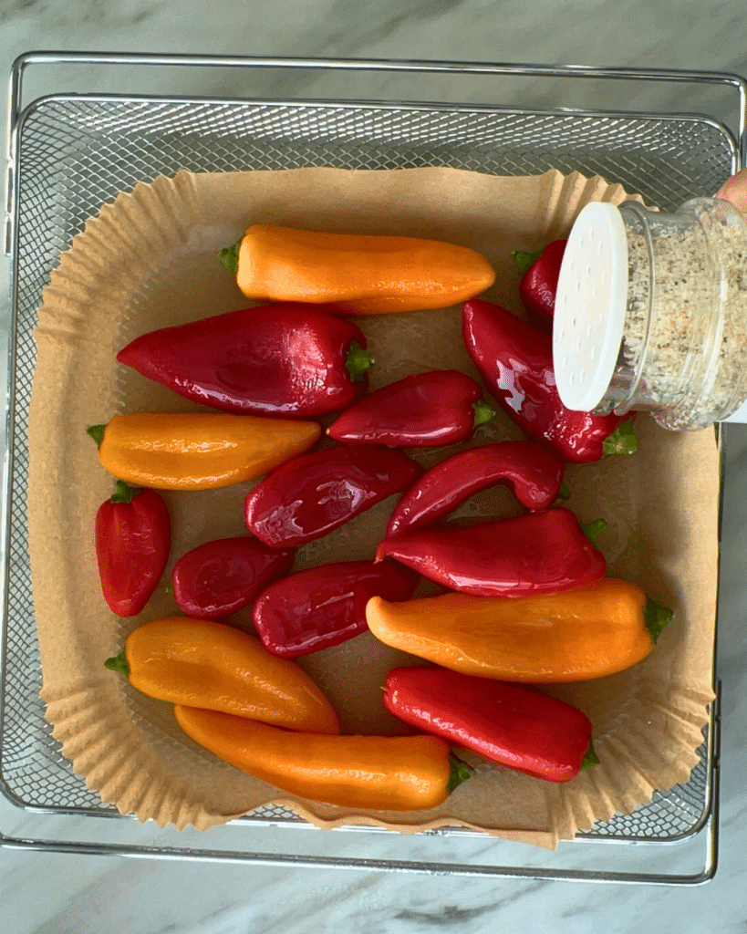 A hand seasons a tray of colorful mini sweet peppers and air fryer baby potatoes on parchment paper before air frying.
