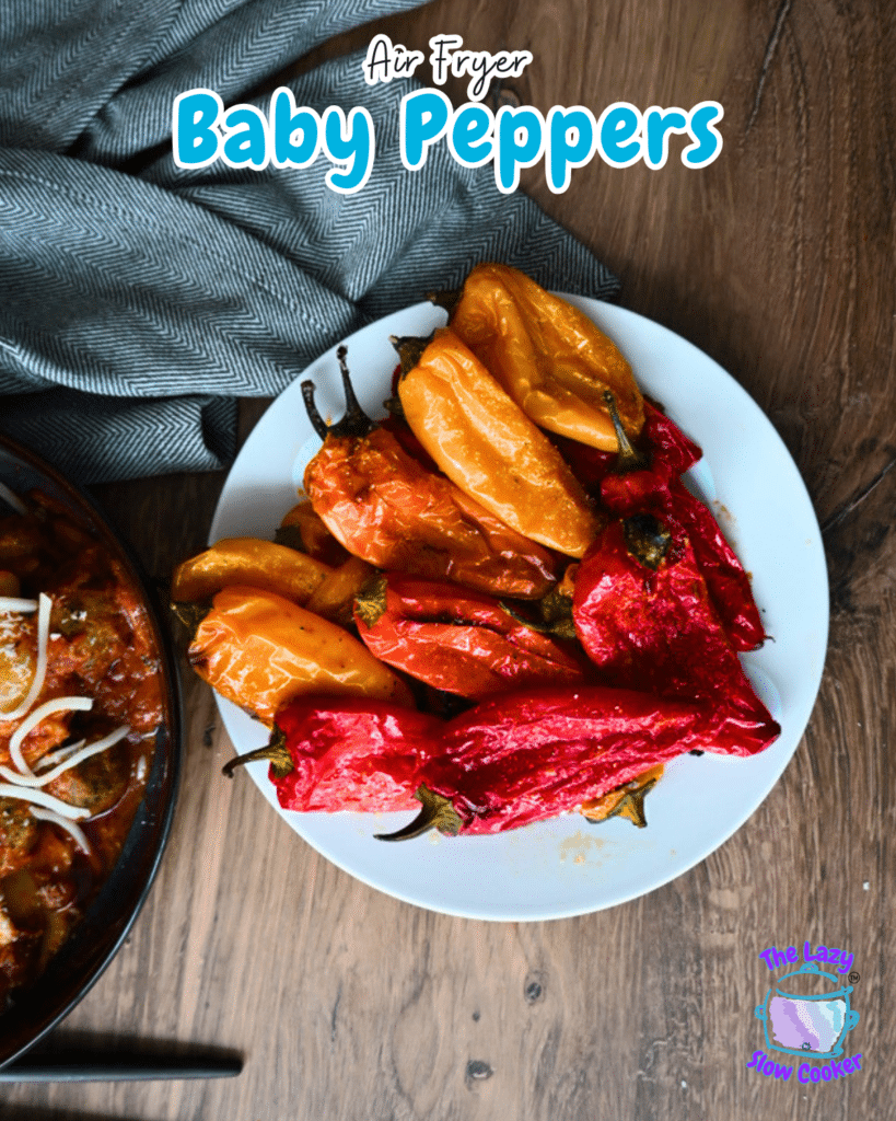 A white plate with roasted baby peppers and potatoes sits on a wooden table beside a blue-gray cloth. Text reads Air Fryer Baby Peppers.