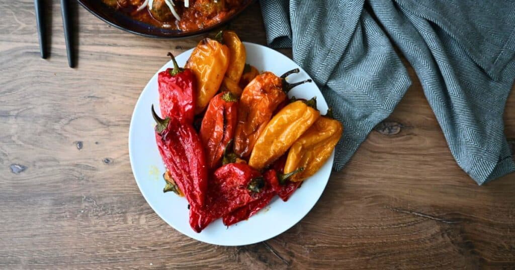 A white plate of roasted red and yellow peppers is on a wooden table next to a blue-gray cloth, with air fryer potatoes in back.