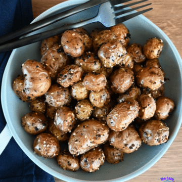 A bowl of golden, seasoned air fryer baby potatoes sits on a wooden table with a black fork, knife, and dark napkin beside it.