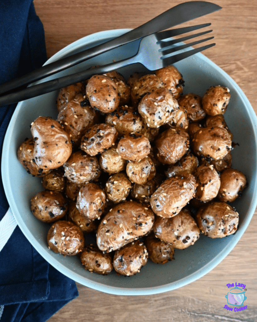 A bowl of golden, seasoned air fryer baby potatoes sits on a wooden table with a black fork, knife, and dark napkin beside it.