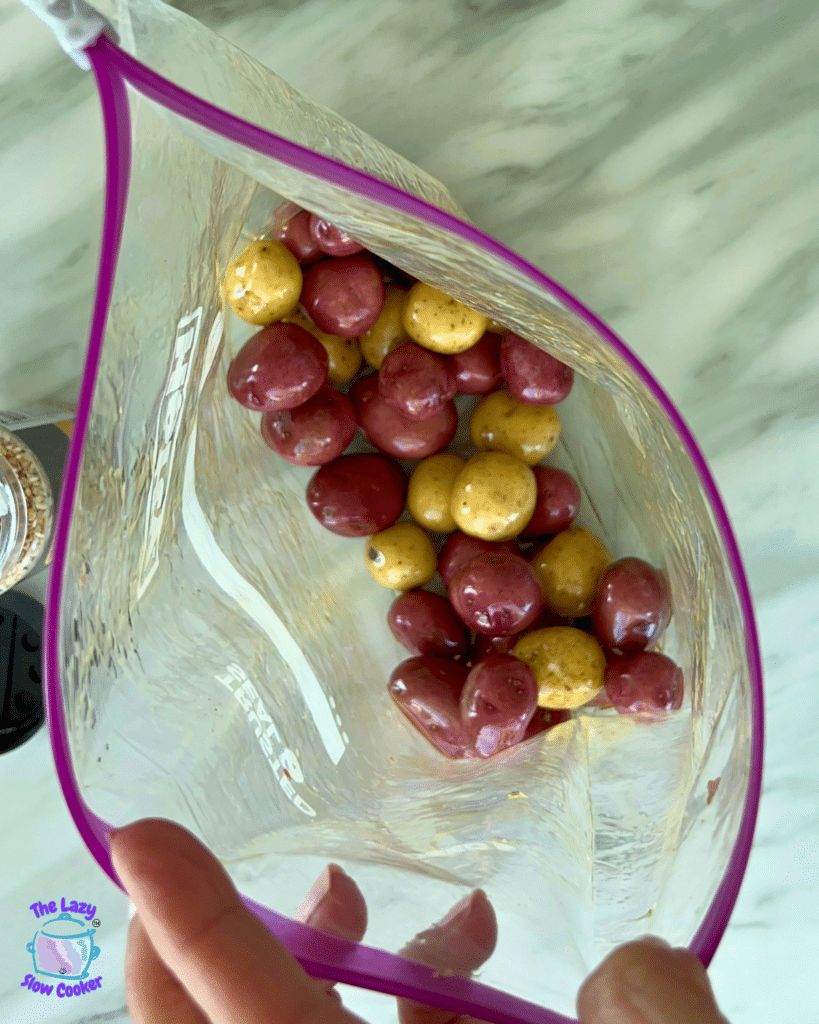 A hand holds a clear, purple-trimmed bag of small red and yellow air fryer potatoes over a light marble surface; The Lazy Slow Cooker logo appears in the corner.