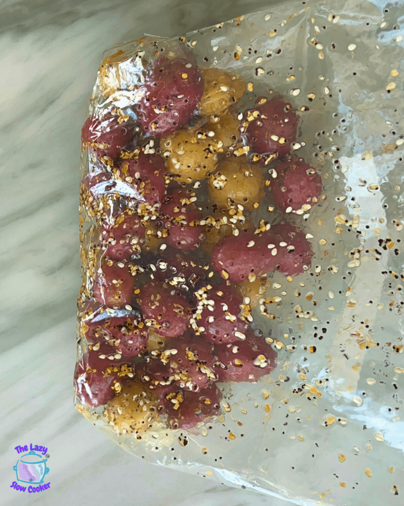 Red and yellow baby potatoes coated in oil and seasoning sit in a plastic bag on marble; The Lazy Slow Cooker logo appears in the corner.