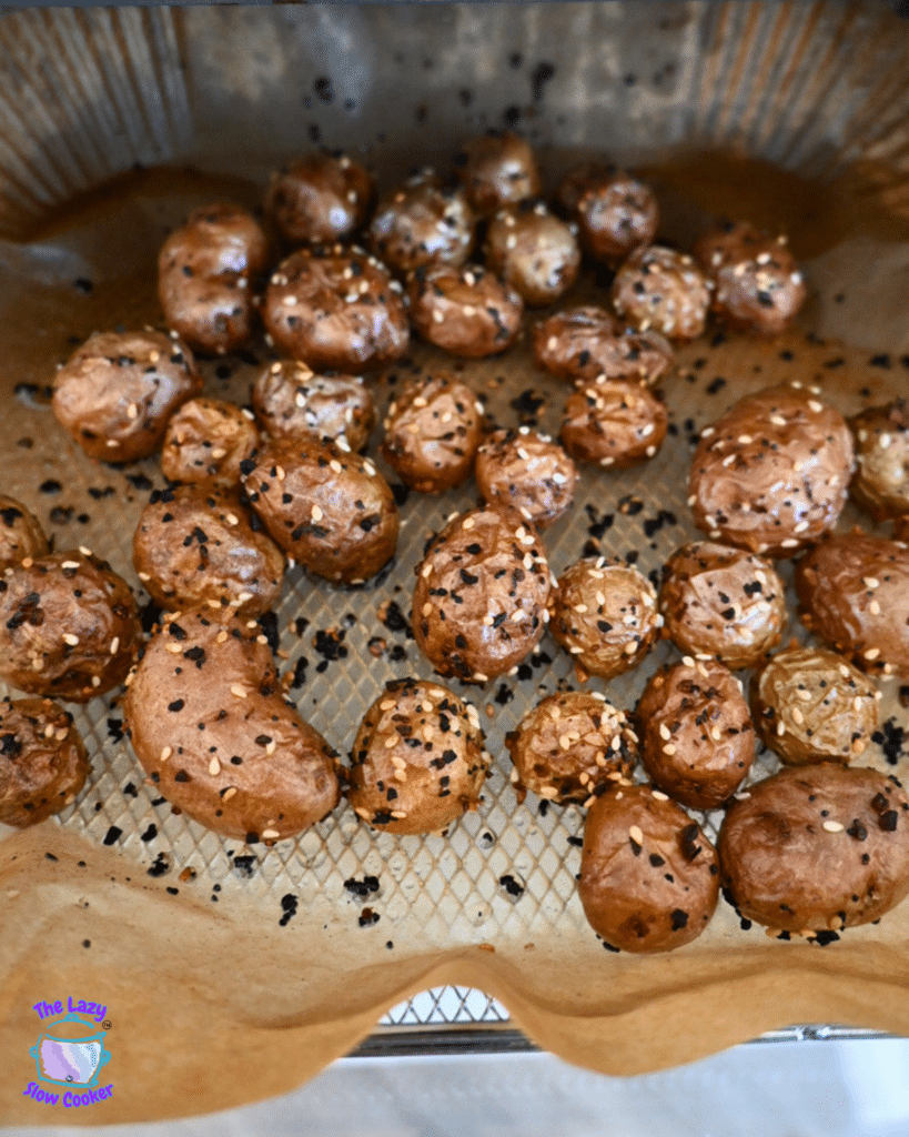 Golden brown baby potatoes cooked in an air fryer sit in a single layer on a parchment-lined tray, topped with seeds and seasoning.