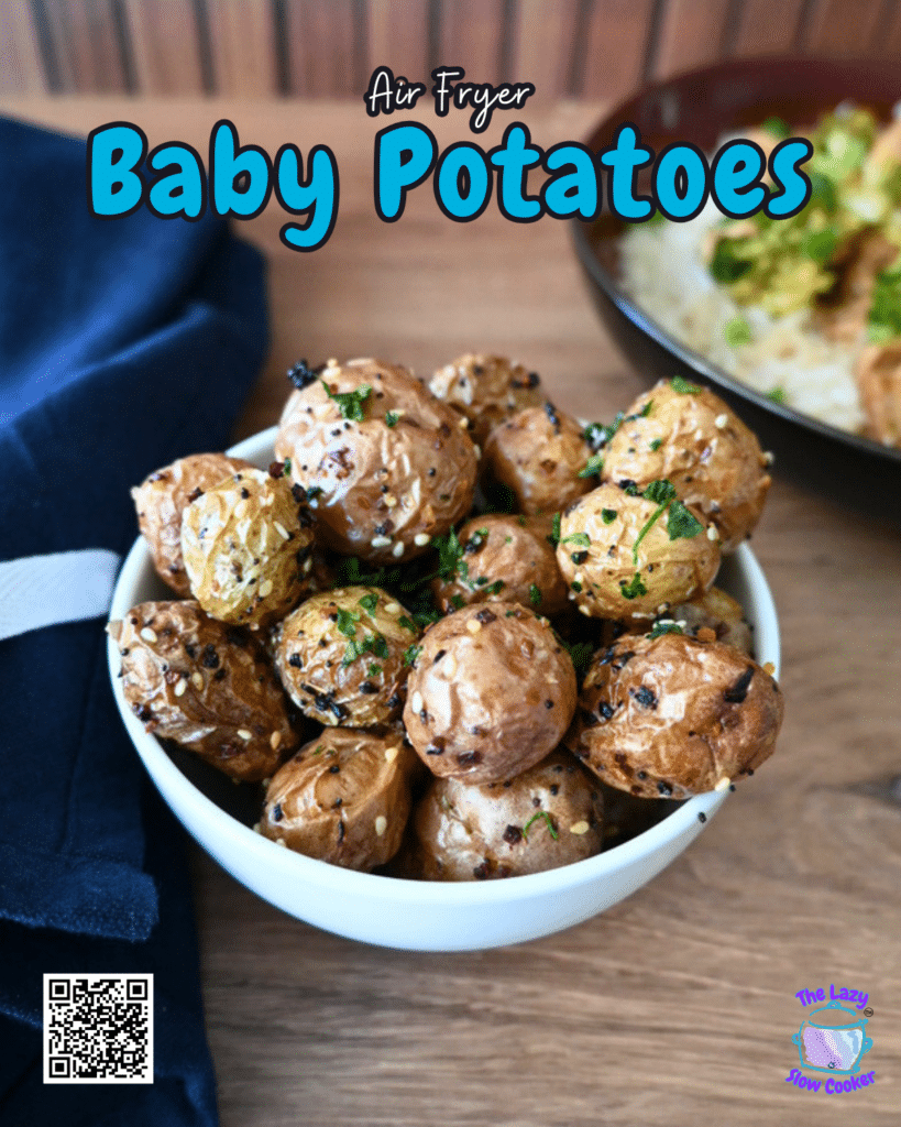A bowl of air fryer baby potatoes topped with herbs and sesame seeds on a wooden table, with a napkin and blurred plate behind.