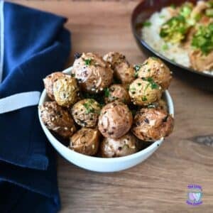 A white bowl of air fryer baby potatoes topped with herbs and sesame seeds sits on a wooden table, napkin and plate in background.