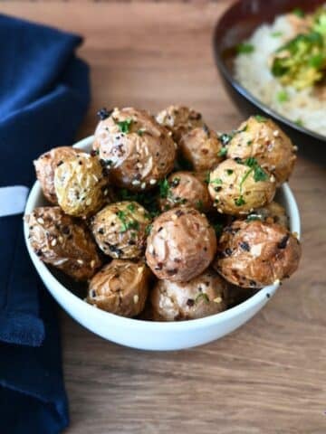 A white bowl of air fryer baby potatoes topped with herbs and sesame seeds sits on a wooden table, napkin and plate in background.