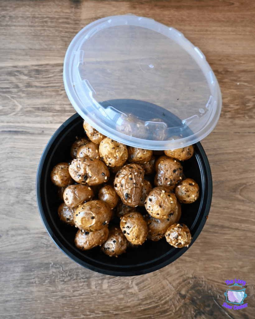 Seasoned, golden air fryer baby potatoes in a black plastic container with a clear lid, resting on a wooden table.
