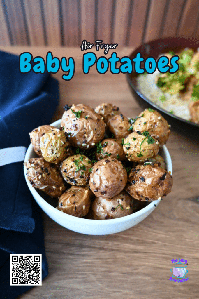 A bowl of air fryer baby potatoes topped with herbs and sesame seeds on a wooden table, with a napkin and blurred plate behind.