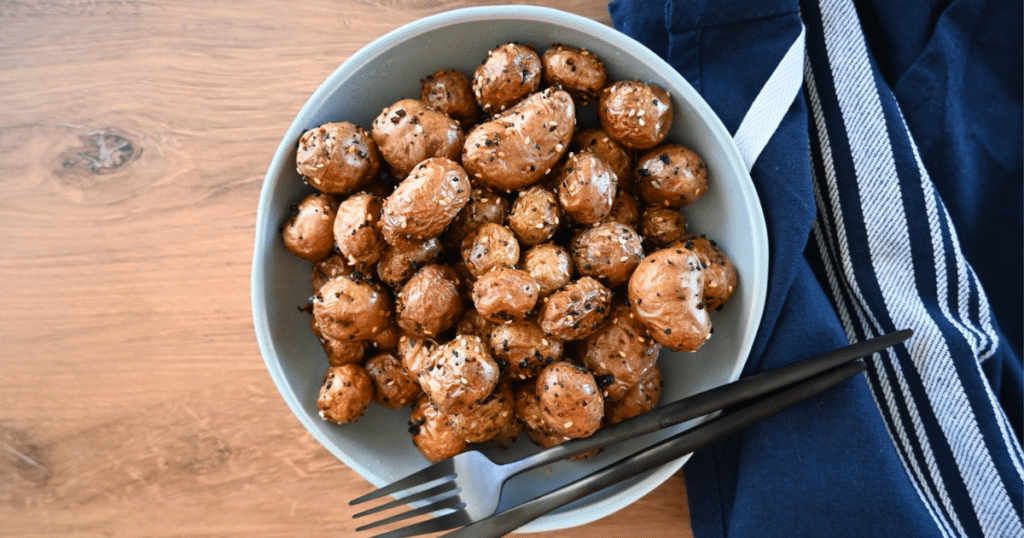 Herbed baby potatoes cooked in an air fryer are served in a bowl on a wooden table with cutlery and a blue-striped napkin nearby.