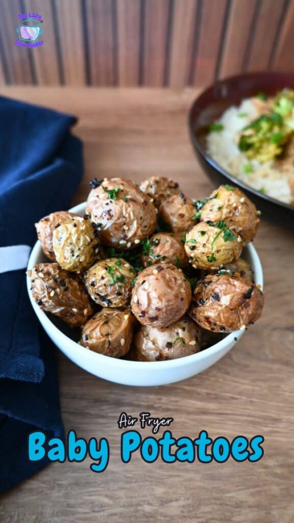 A white bowl of golden air-fried baby potatoes with herbs and sesame seeds sits on a wooden table beside a navy napkin.
