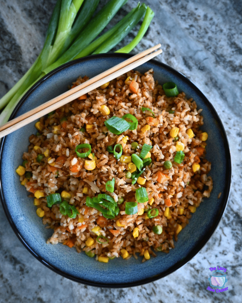 A bowl of vegetable fried rice topped with green onions and chopsticks rests on a marble surface, with fresh onions beside it.