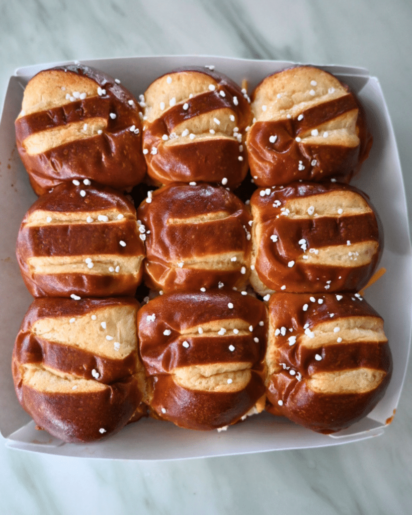 Nine shiny brown pretzel rolls, each with coarse salt and three slashes, sit in a white tray—ideal for slow cooker BBQ sliders.