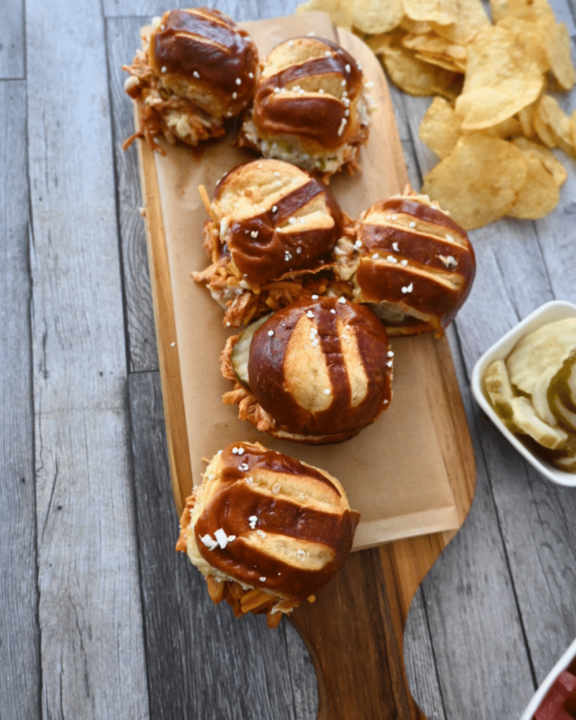 Six BBQ chicken pretzel sliders on a wood board with parchment, served with potato chips and a bowl of pickles on a gray table.