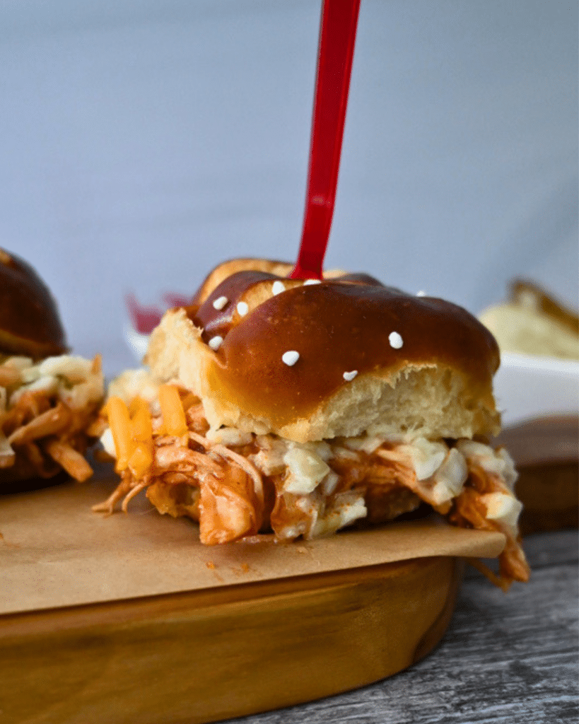 Close-up of slow cooker BBQ chicken sliders with pulled chicken, melted cheese, and coleslaw on pretzel buns, held with red picks.