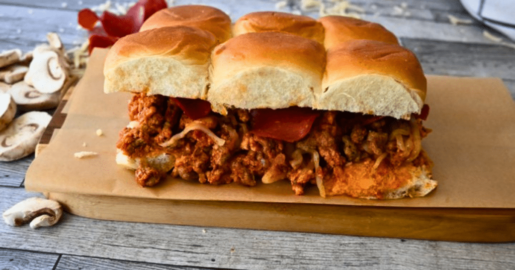 Six connected pizza sloppy Joe sliders filled with ground meat, melted cheese, and pepperoni on a wooden board, mushrooms scattered beside.