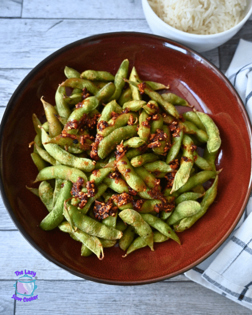 A red bowl of air-fried edamame with spicy garlic seasoning sits on a wooden table beside white rice and a striped napkin.