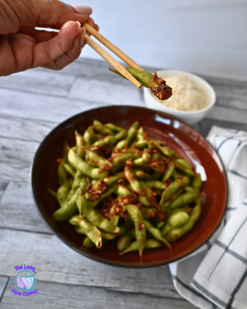 A hand uses chopsticks to pick up a spicy air fryer edamame pod from a bowl, with rice and a striped napkin on the wooden table.
