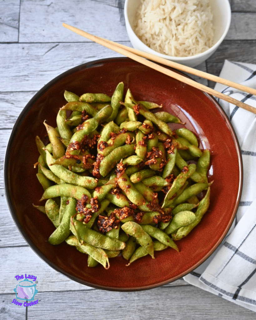 Spicy air-fried edamame with chili sauce on a brown plate beside white rice and chopsticks on a striped napkin and wood table.