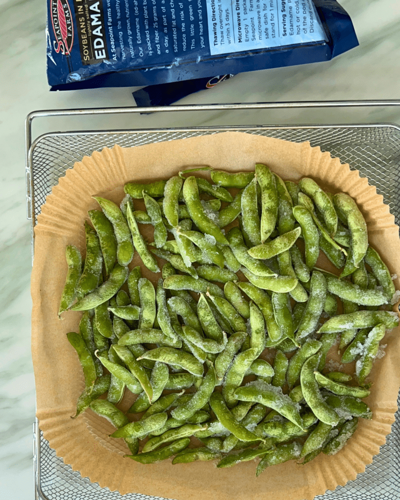 A parchment-lined basket holds cooked edamame pods from the air fryer, with an open bag of frozen edamame behind on a light counter.