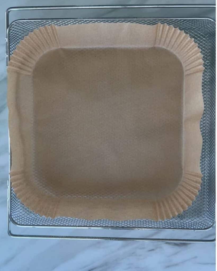 A square piece of brown parchment paper lines a wire mesh air fryer basket, sitting on a white marble countertop.