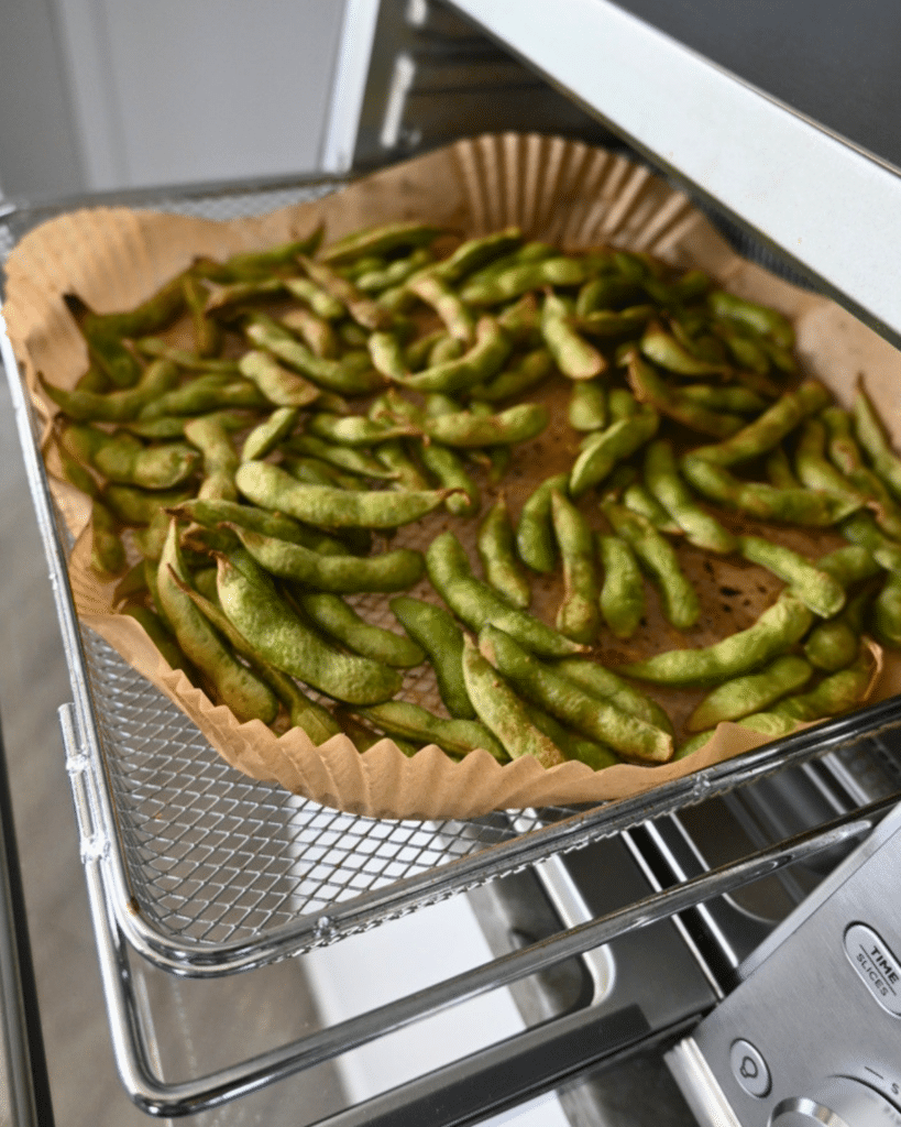 A parchment-lined tray with crisp green edamame pods sits in an open air fryer oven, just finished cooking with the air fryer method.