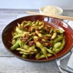 A bowl of air-fried spicy edamame with chili flakes and garlic sits on a wooden table, rice and chopsticks in the background.
