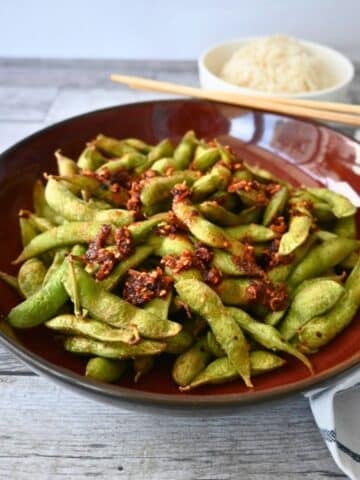 A bowl of air-fried spicy edamame with chili flakes and garlic sits on a wooden table, rice and chopsticks in the background.