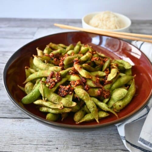A bowl of air-fried spicy edamame with chili flakes and garlic sits on a wooden table, rice and chopsticks in the background.