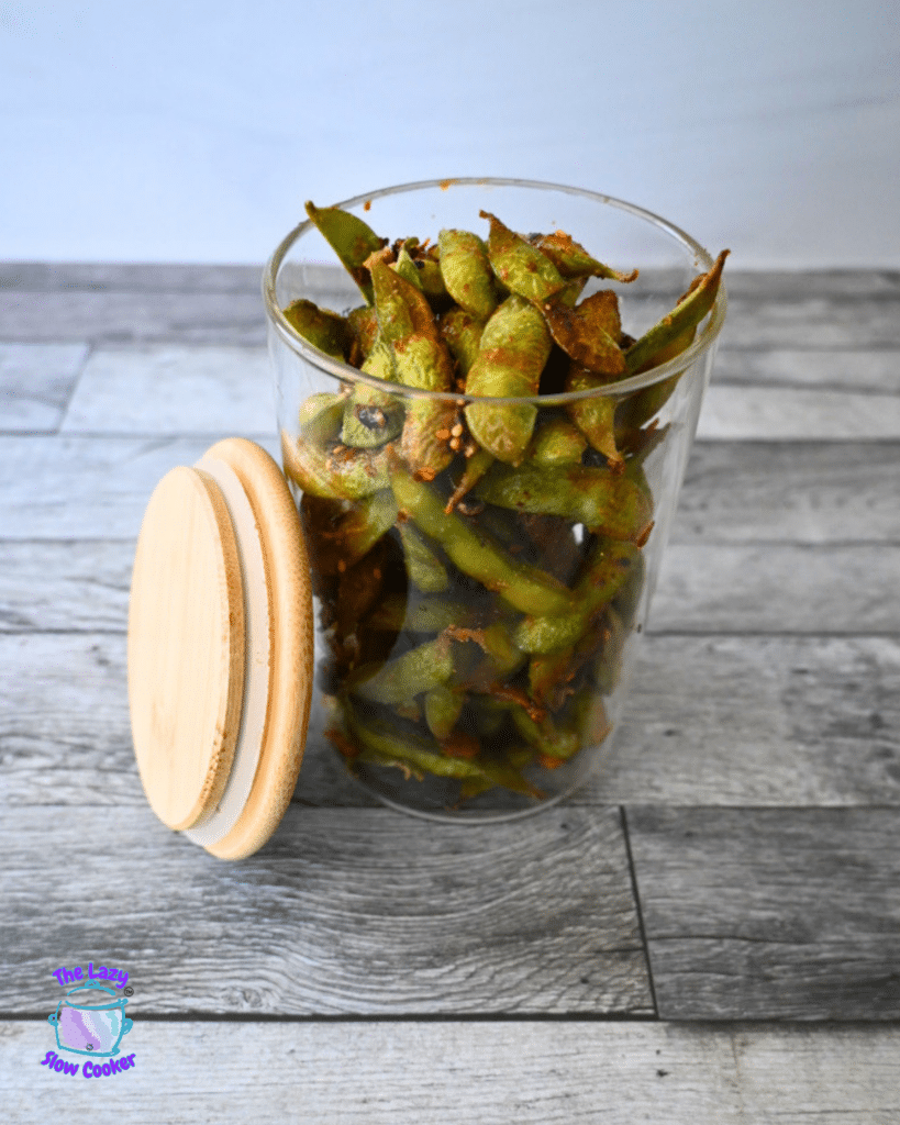 A glass jar of air-fried seasoned edamame pods sits open on a gray wood surface, lid nearby, with The Lazy Slow Cooker logo in view.