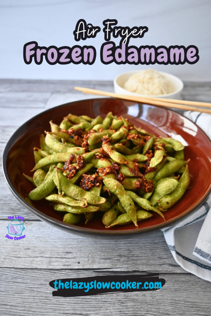 A brown bowl of air fryer edamame with chili sauce sits on a gray table, next to chopsticks and a small bowl of rice. Text reads Air Fryer Frozen Edamame.