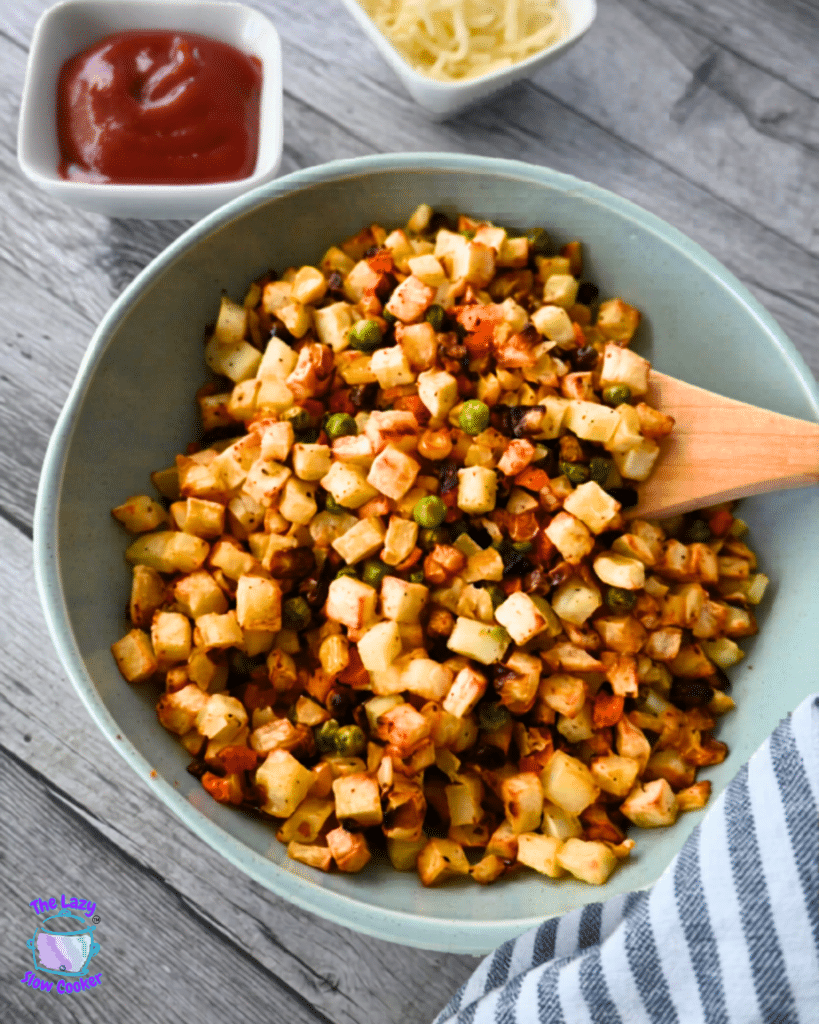 A bowl of diced potatoes, carrots, and peas with a wooden spoon sits by ketchup and grated cheese