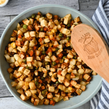 A bowl of diced potatoes, carrots, peas, and corn sits on a striped cloth with a wooden spoon labeled The Lazy Slow Cooker.