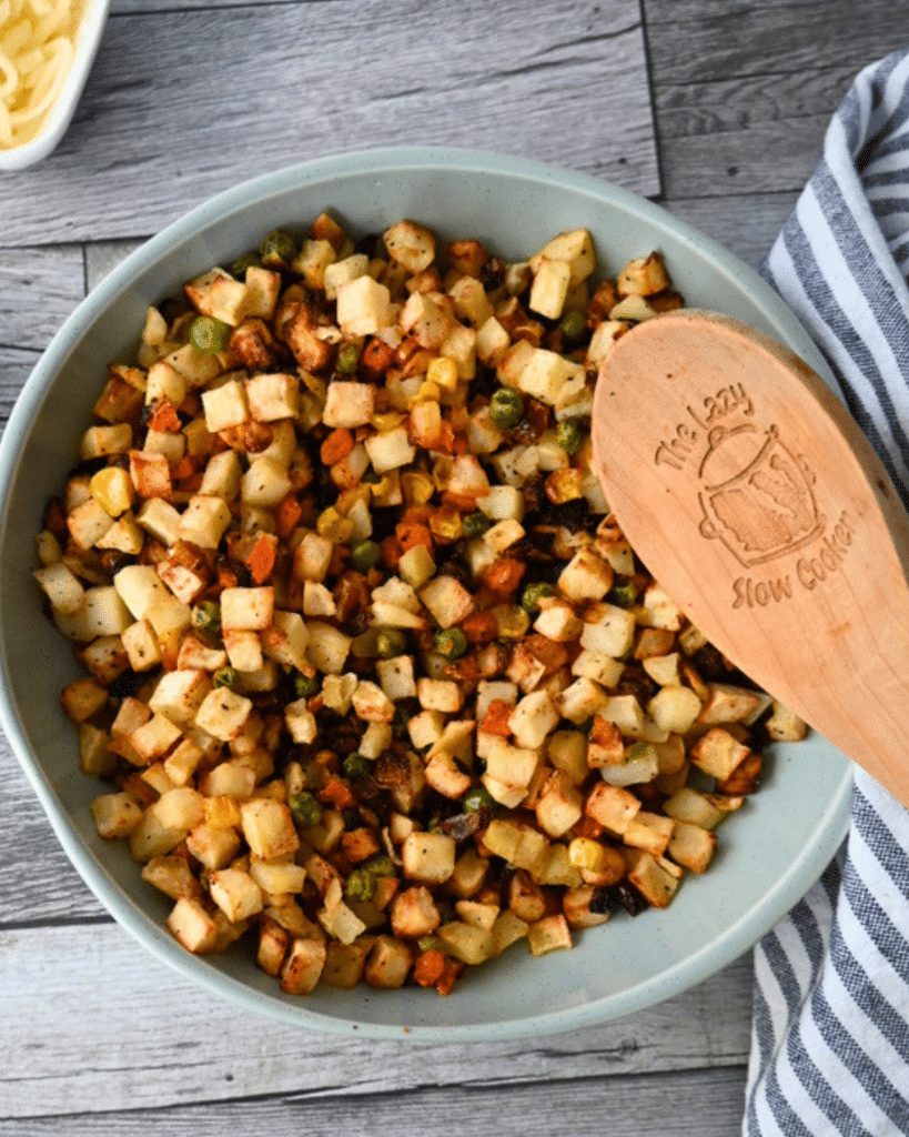 A bowl of diced potatoes, carrots, peas, and corn sits on a striped cloth with a wooden spoon labeled The Lazy Slow Cooker.