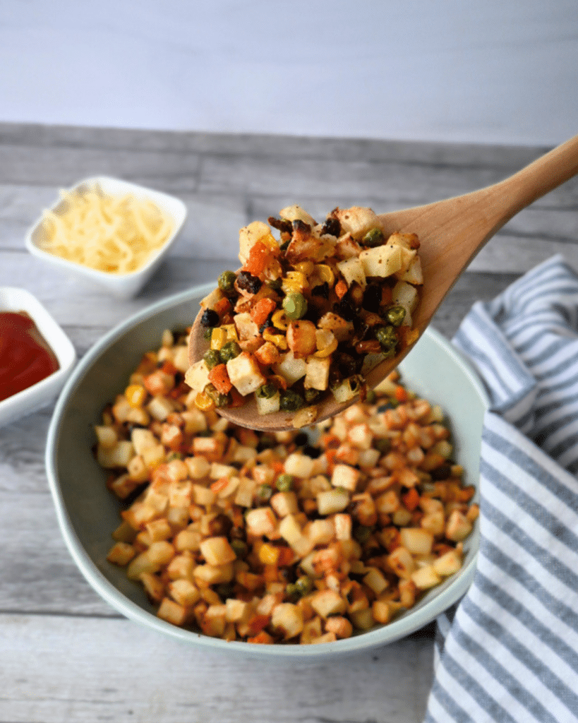 A wooden spoon lifts diced, golden and crispy potatoes and veggies over a bowl of the same, with grated cheese and ketchup nearby