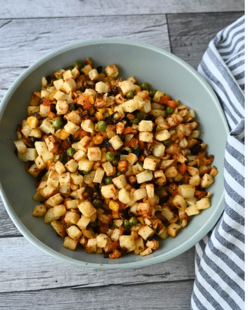 A bowl of diced potatoes, peas, and carrots with spices sits on a wooden table next to a striped cloth, served with BBQ chicken sliders.