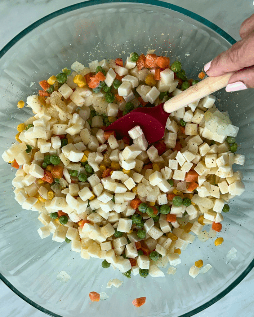 A hand uses a red spatula to stir diced potatoes, carrots, peas, and corn in a glass bowl in preparation for the air fryer
