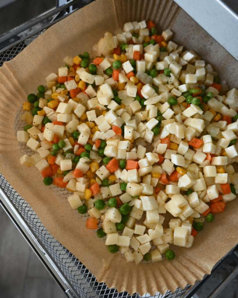A mix of chopped peas, carrots, corn, and potatoes lies on parchment in an air fryer basket