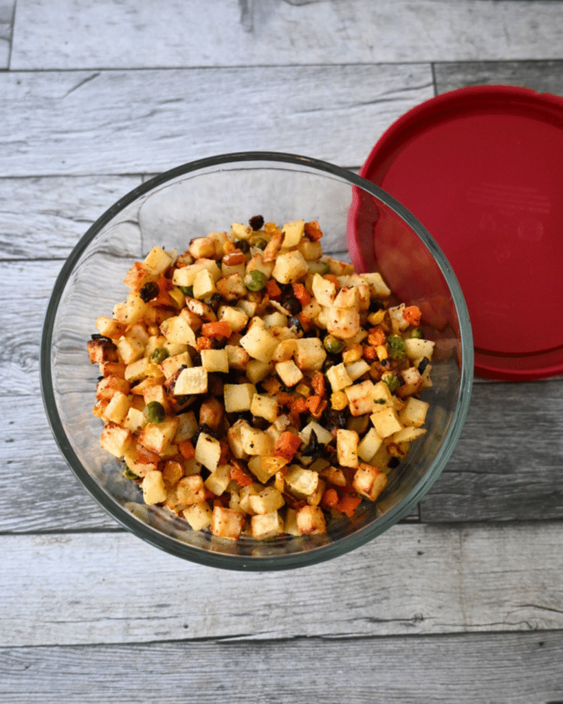 A glass bowl of leftover air fried potatoes and mixed vegetable salad sits on a wood surface with a red lid nearby ready for storing in the refrigerator.