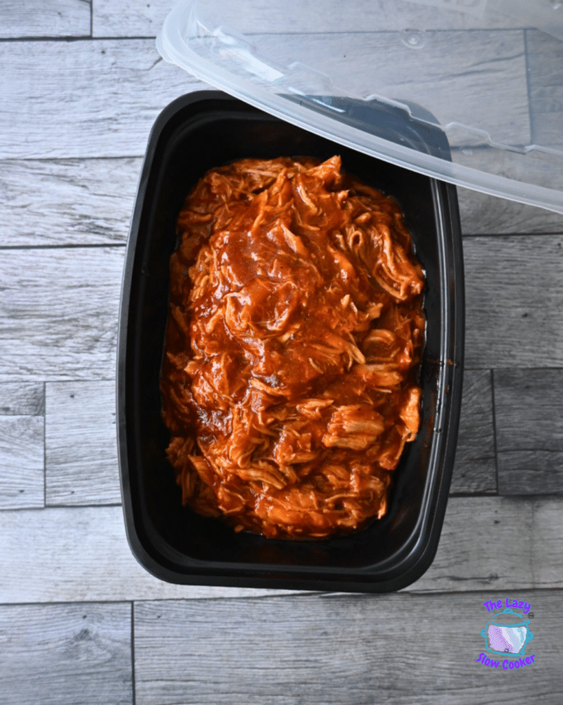 Shredded BBQ chicken in a black slow cooker with the lid slightly open, resting on a gray wood surface.