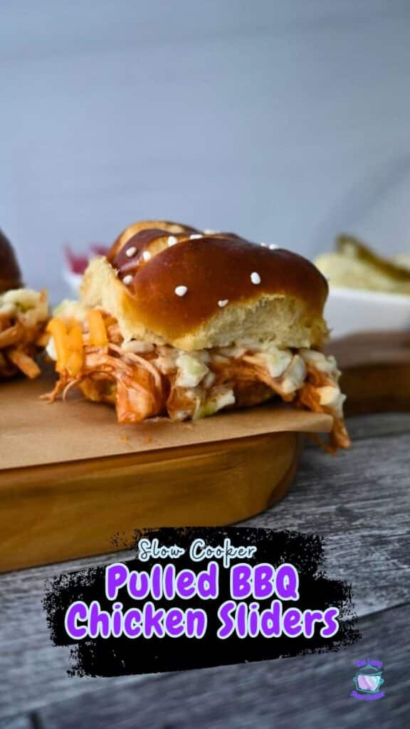 A BBQ chicken slider with melted cheese and coleslaw on a pretzel bun rests on a wooden board. Text says Slow Cooker BBQ Chicken Sliders.