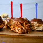 Three BBQ chicken sliders made in a slow cooker sit on a wooden board, topped with cheese and bright skewers; flowers blurred behind.