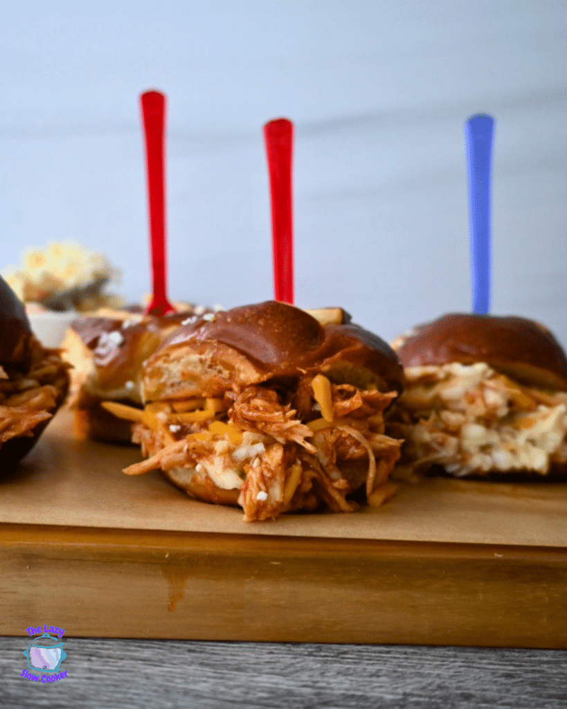 Three BBQ chicken sliders made in a slow cooker sit on a wooden board, topped with cheese and bright skewers; flowers blurred behind.