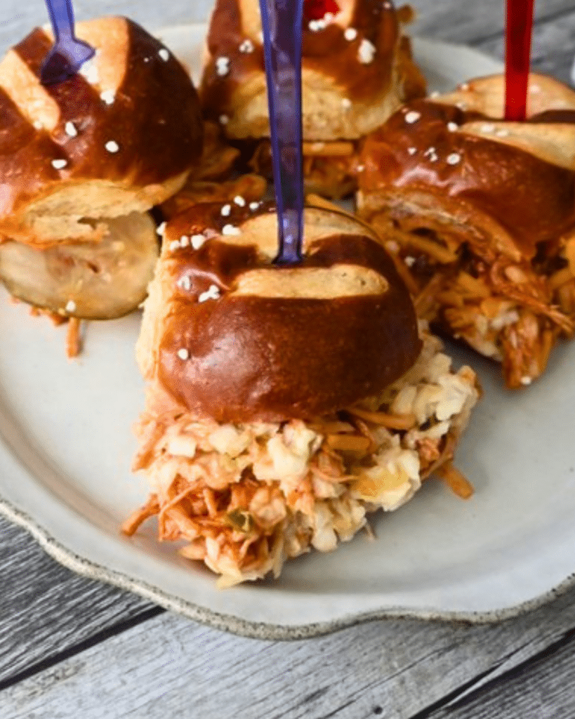 Four slow cooker BBQ chicken sliders with shredded meat and coleslaw in pretzel buns sit on a white plate on rustic wood.