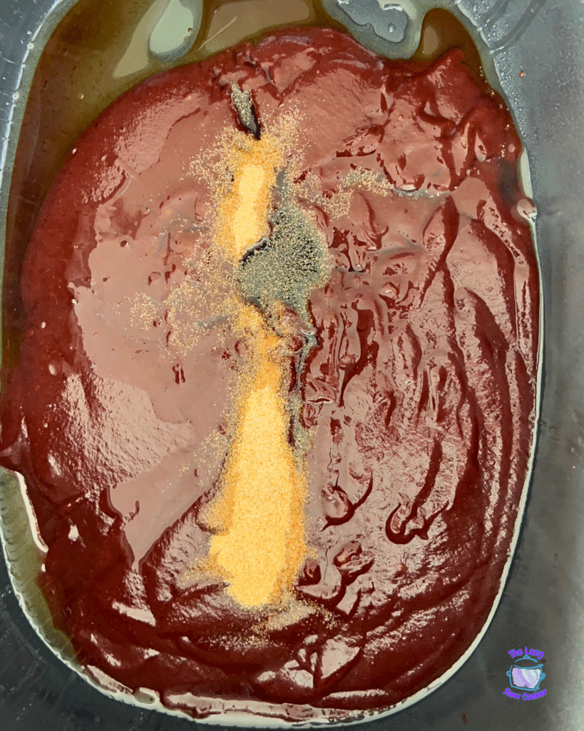 A slow cooker filled with dark red barbecue sauce, topped with small heaps of yellow and light brown spices for shredded BBQ chicken sliders.
